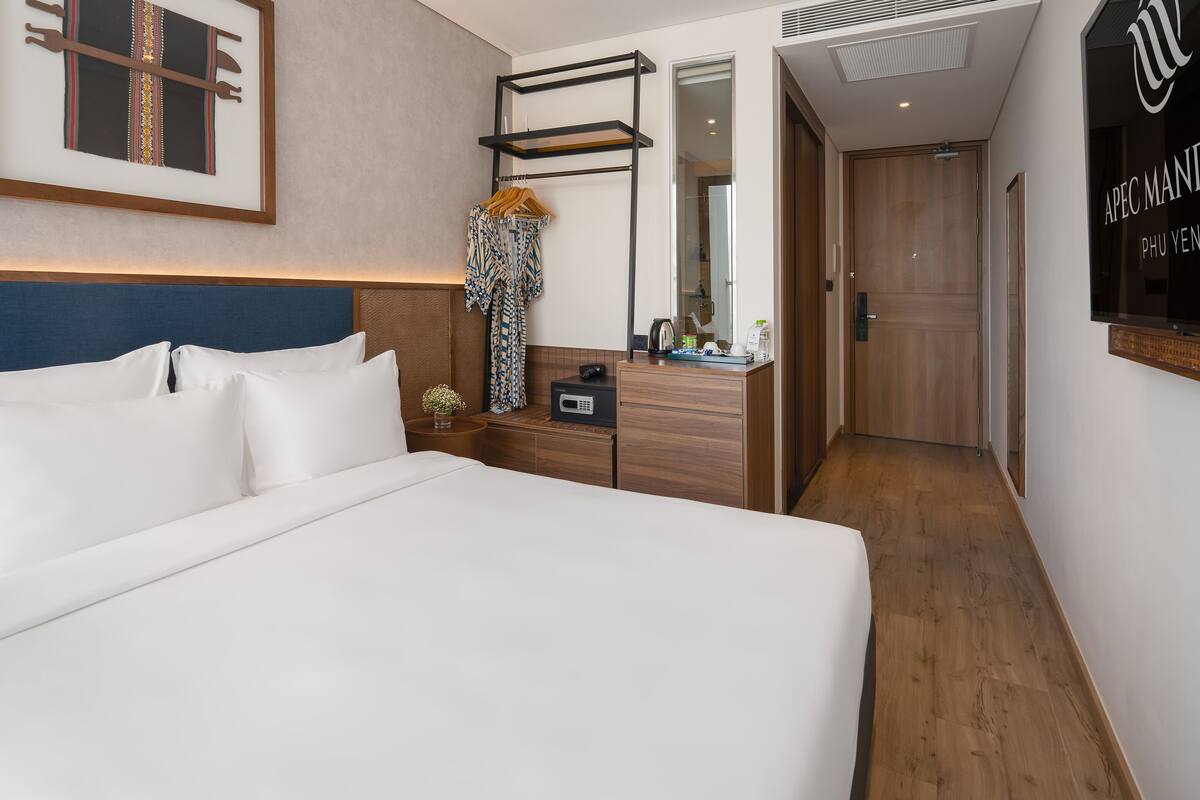 A hotel room with a large bed, a wooden headboard, a bedside table with a lamp, a mirror, and a framed picture on the wall.