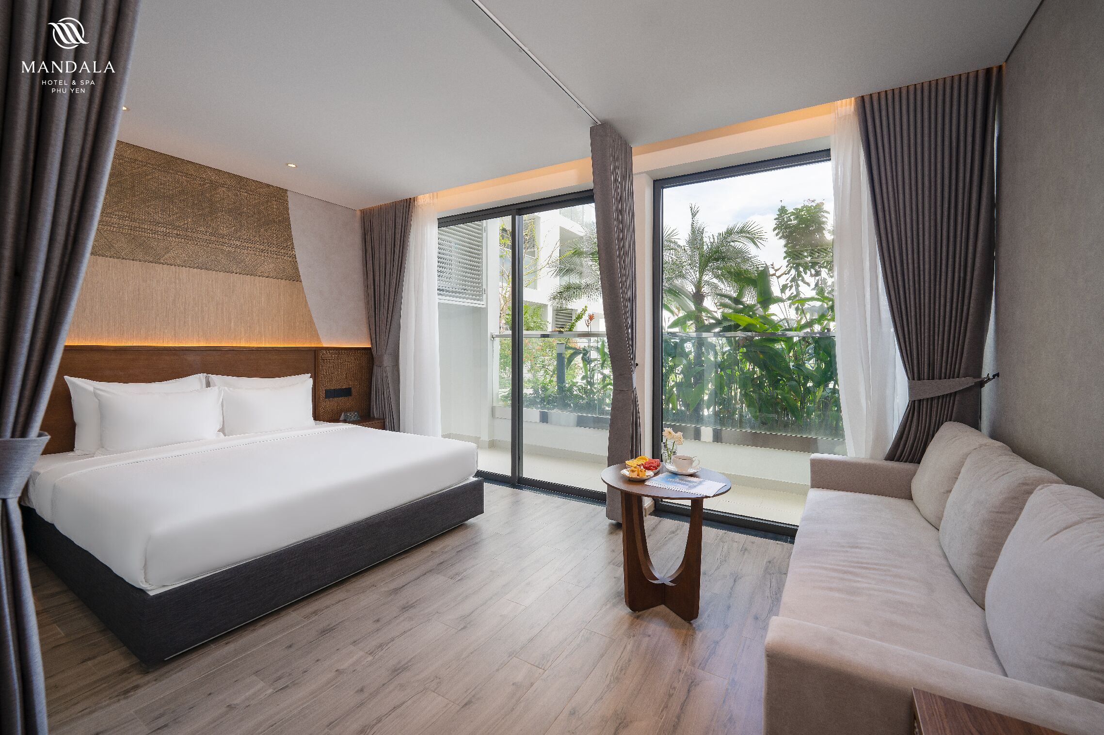 Una habitación de hotel con una cama grande, un sofá, una mesita con un frutero y vistas a zonas verdes a través de amplios ventanales.