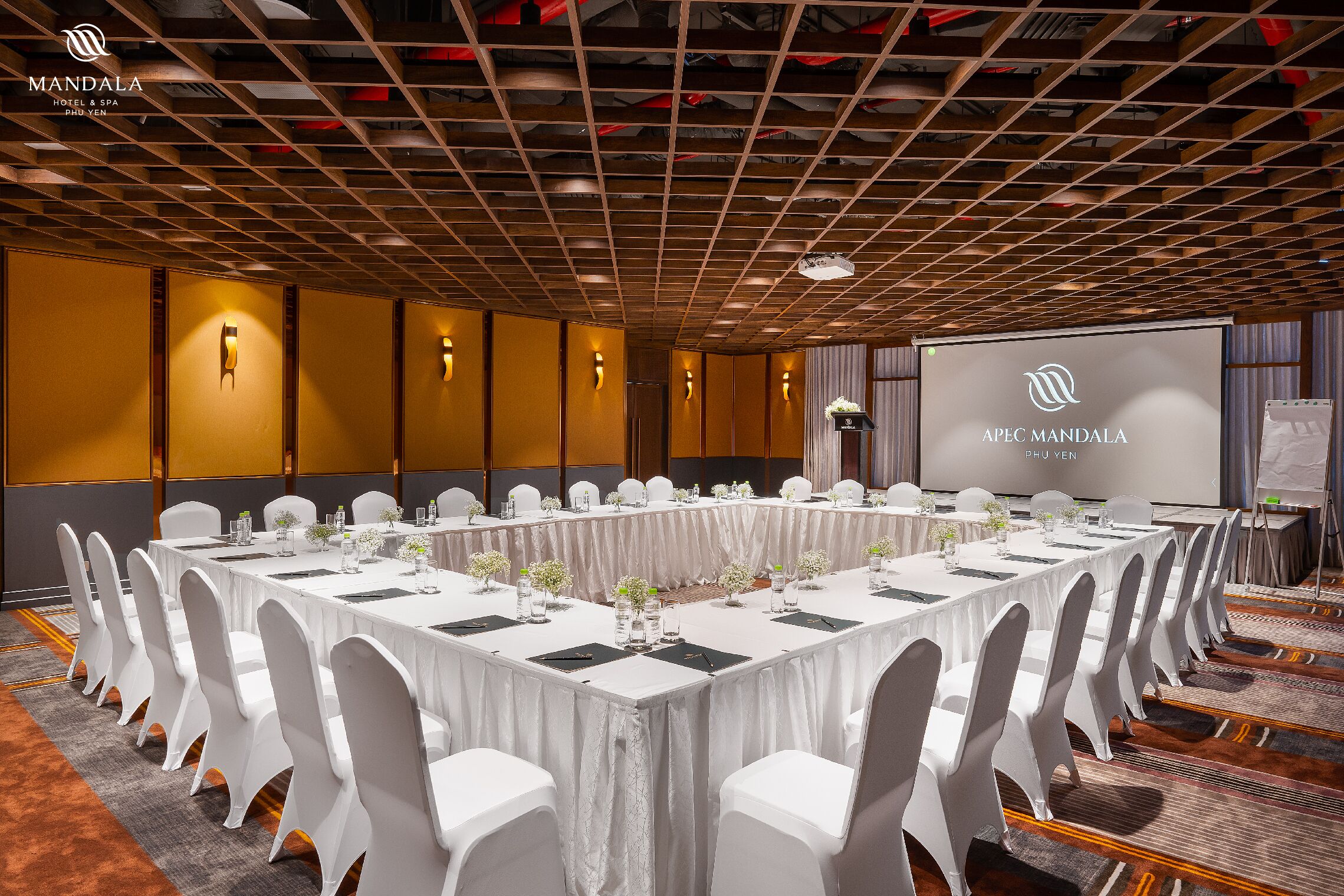 A conference room with a long table, white chairs, and a projector screen displaying "APEC Mandala PHU VEN".
