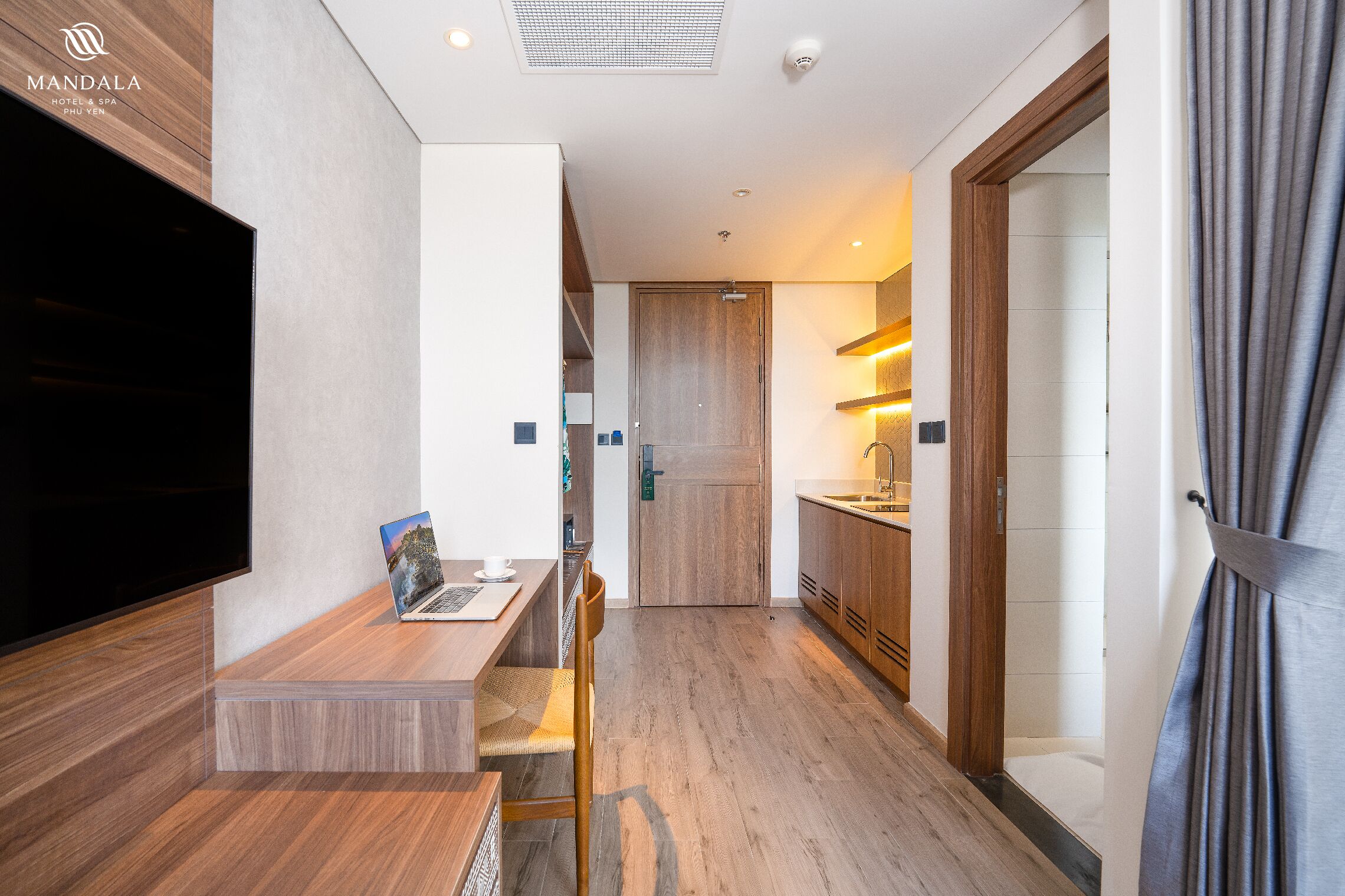 A modern hotel room with a wooden floor, a flat-screen TV, a laptop on a wooden table, and a shower area visible through a glass door.