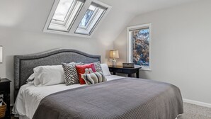 Ein Schlafzimmer mit schrägverdeckter Decke, einem Bett mit grauem Kopfteil, einem Nachttisch, einer Lampe und einem Fenster mit Blick auf Bäume.