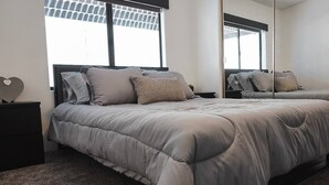 Een net opgemaakt bed met een grijze deken en kussens, een nachtkastje van donker hout, en een grote spiegel die de kamer weerspiegelt.
