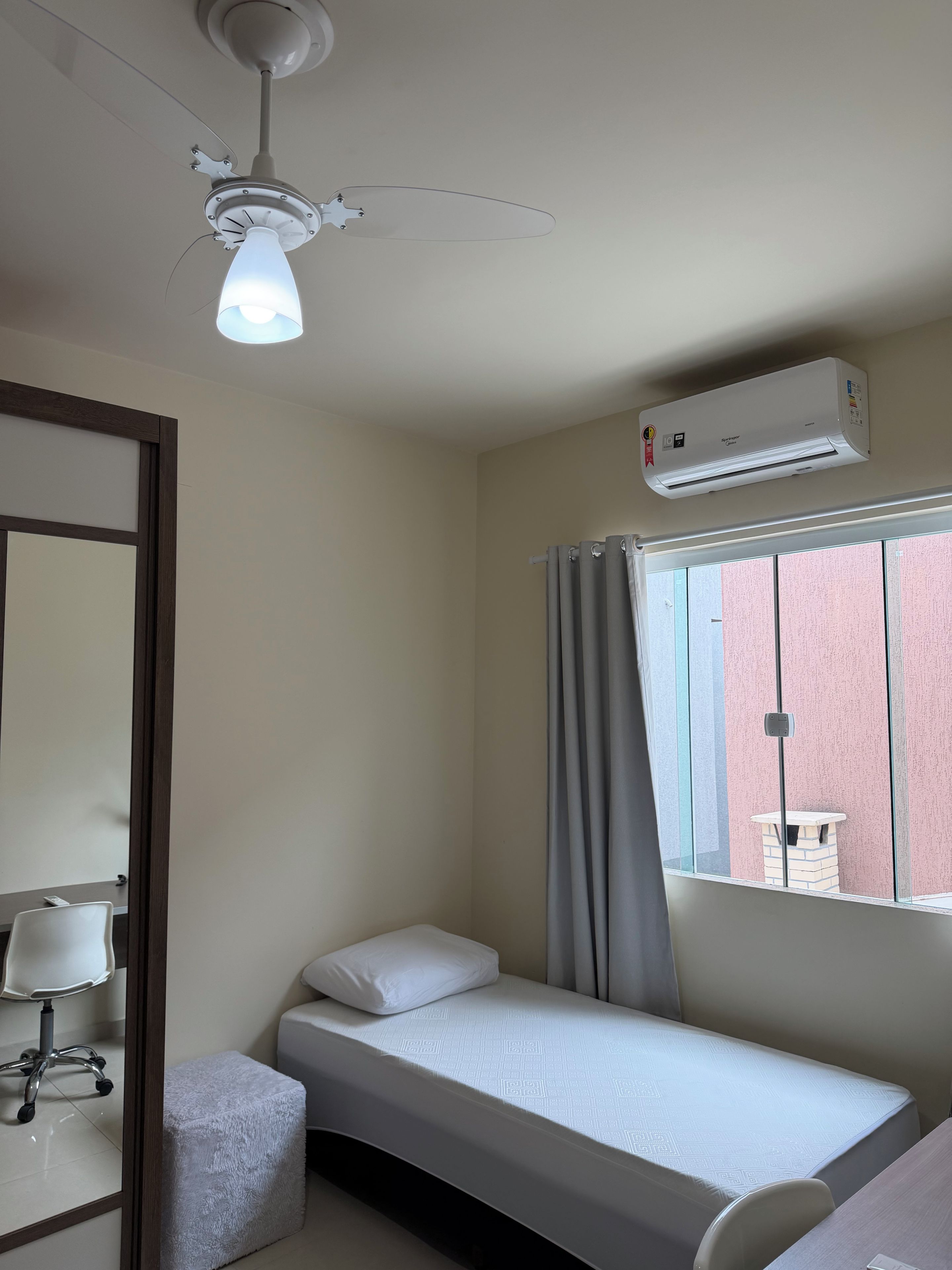 A single bed with a white mattress and pillow, a bedside table, a chair, a ceiling fan, and an air conditioning unit.