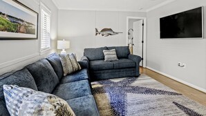 A living room with a sectional sofa, a ceiling fan, a mounted fish decoration, and a large TV on the wall. - The Saltwater Cottage-Golf Cart & Linens Included in Ocean Lakes (Myrtle Beach)