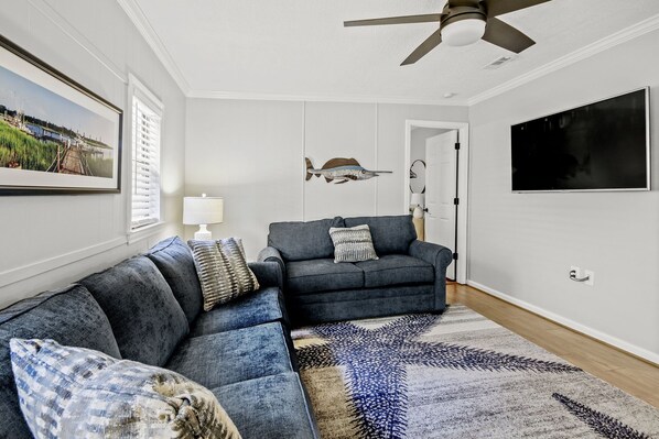 A living room with a sectional sofa, a ceiling fan, a mounted fish decoration, and a large TV on the wall.