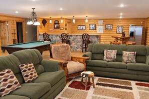 A cozy living room with a pool table, green sofas, a wooden chair, and a stone accent wall.