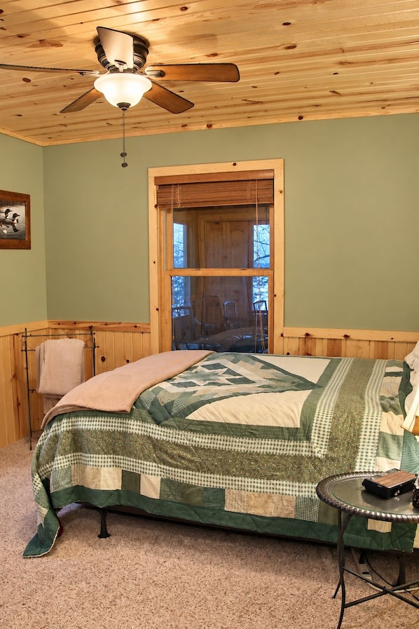 A bedroom with a wooden ceiling, a bed with a quilt, a window with blinds, and a ceiling fan.