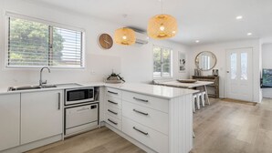 A modern kitchen with white cabinetry, a built-in oven, a microwave, and a sink. There are pendant lights, a clock, and a large window with blinds. - Our Little White House -74  Padman Crescent, Middleton (Middleton)