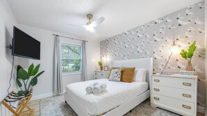 Quarto com cama, mesa de cabeceira, televisão, ventoinha no teto, janela com cortinas e parede com desenho de folhas.