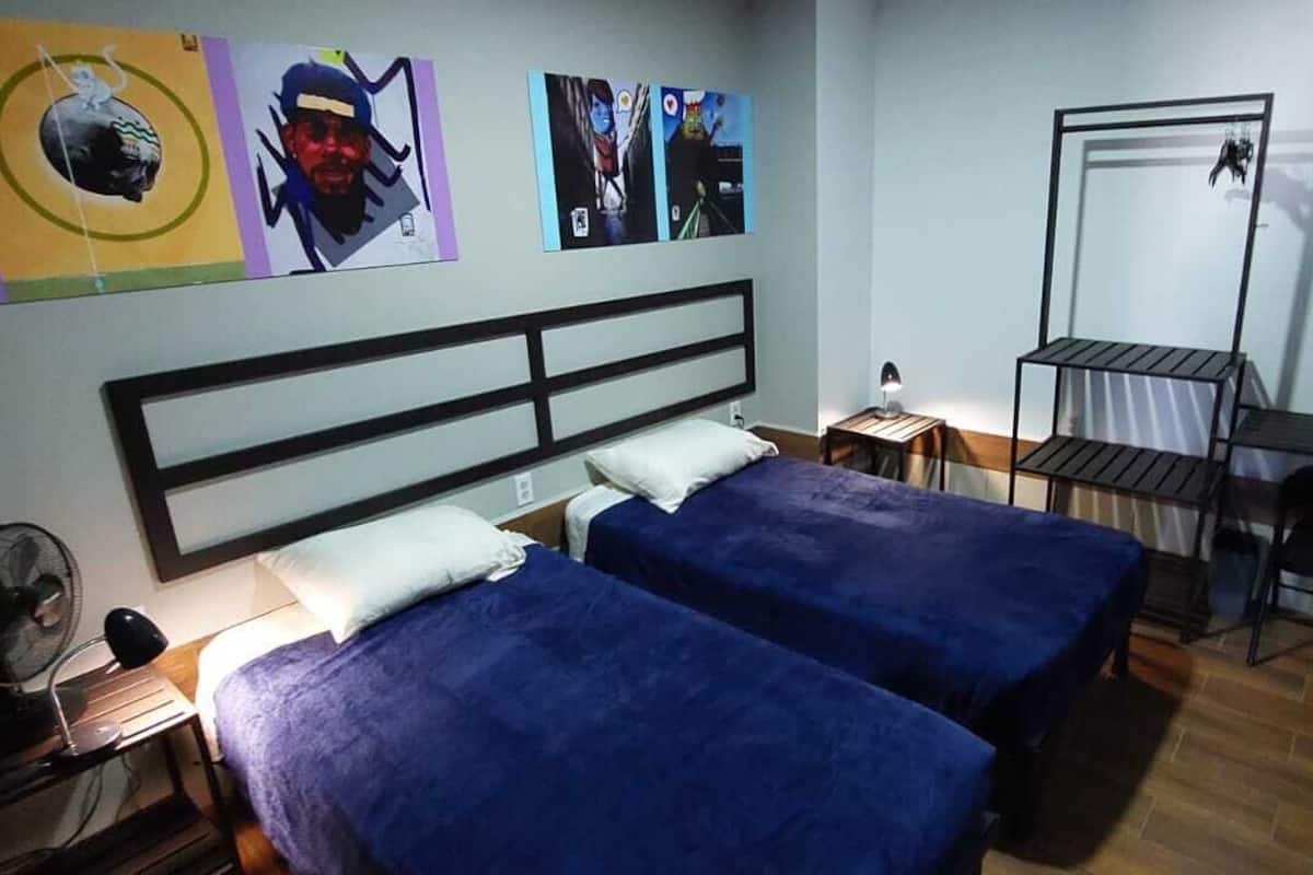 Une chambre avec deux lits, un bureau, une chaise, un ventilateur et des œuvres d’art accrochées au mur.