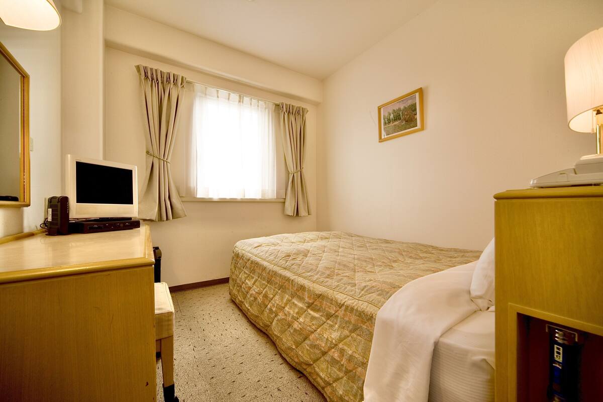 Una habitación de hotel con cama, escritorio, computadora, lámpara y ventana con cortinas.