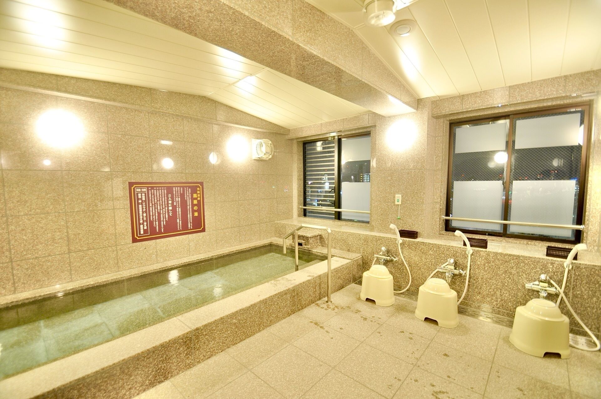 A public restroom with a large bathtub, handwashing stations, and a window with blinds.
