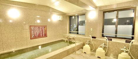 A public restroom with a large bathtub, handwashing stations, and a window with blinds.
