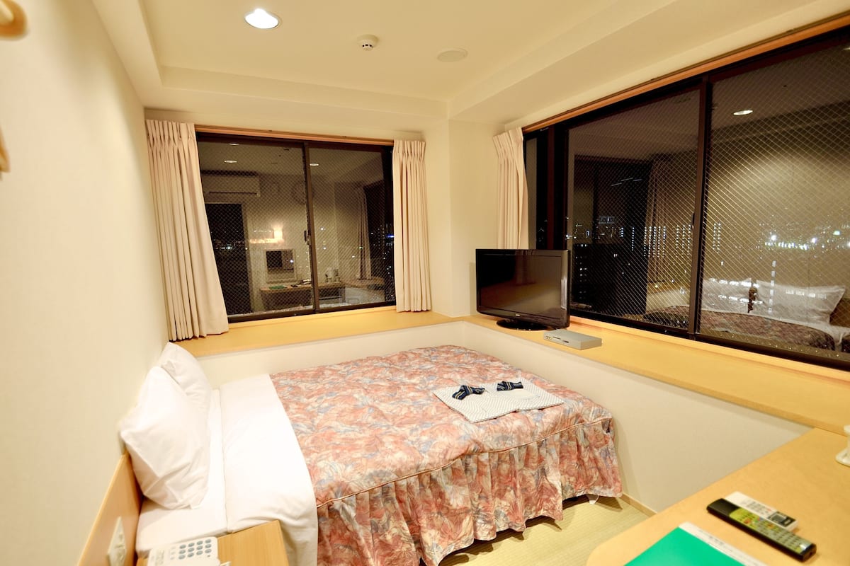 Ein Hotelzimmer mit Bett, Fernseher und großen Fenstern mit Blick auf die Stadt.