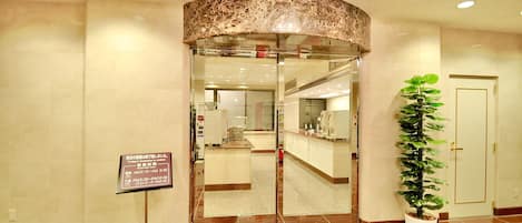 A hotel entrance with a reception desk, a signboard with operating hours, and a potted plant.