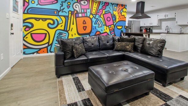 A modern living room with a black leather sectional sofa, a matching ottoman, and a colorful abstract mural on the wall.