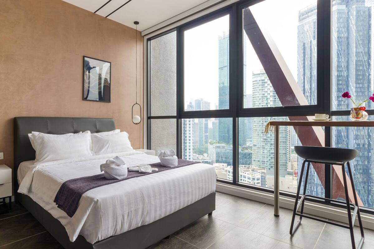 Habitación de hotel moderna con una cama grande, un escritorio con silla y vistas a la ciudad a través de amplios ventanales.