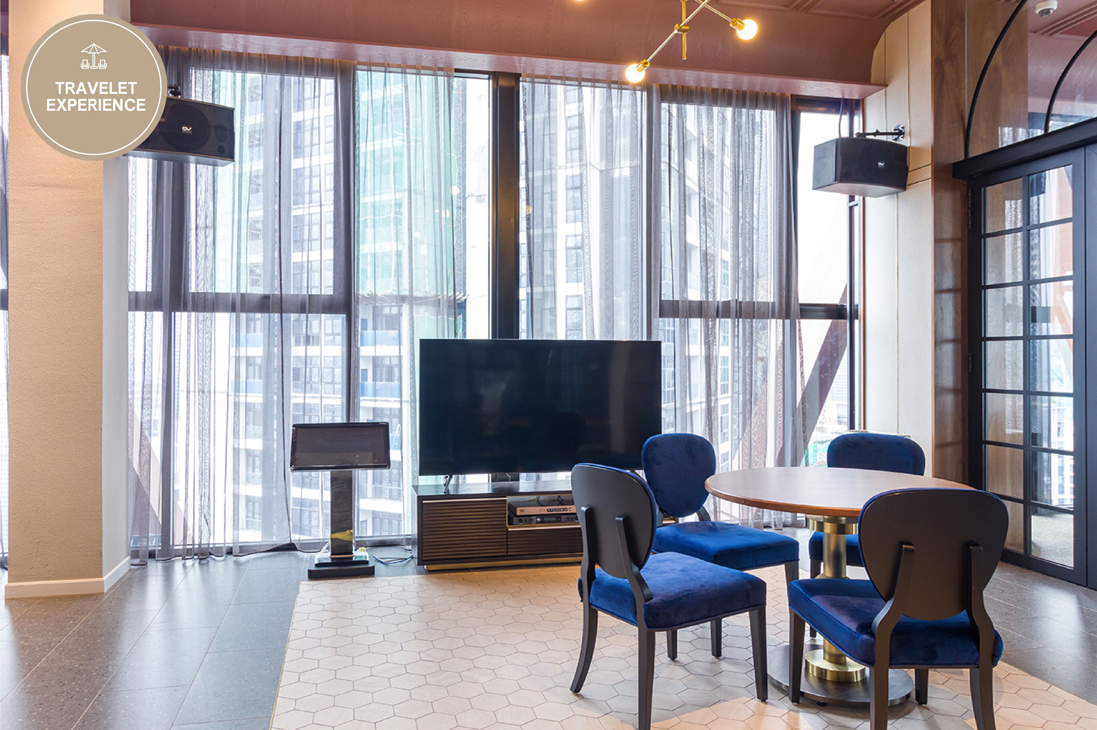 A modern hotel room with a large window, a flat-screen TV, a dining table with chairs, and a speaker on the wall.