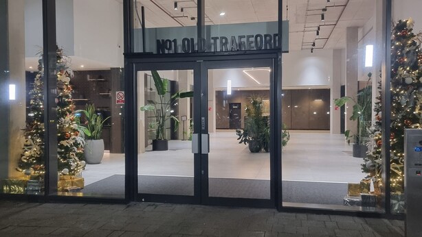 Ein moderner Gebäudeeingang mit Glastüren, einem Schild mit der Aufschrift "No1 Old Trafford" und weihnachtlicher Dekoration.
