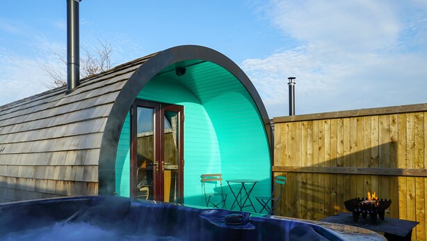Ein gemütlicher Bereich mit Whirlpool im Freien, mit einer Holzterrasse, einer türkisblauen Bogenstruktur und einem Holzzaun im Hintergrund.