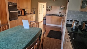 A kitchen with wooden cabinets, a microwave, a coffee maker, and a kettle. There is a table with a laptop and chairs.