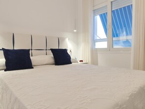 En pænt redt seng med hvid dyne og to mørke puder, et natbord med en rød genstand, og et vindue med lodrette persienner.