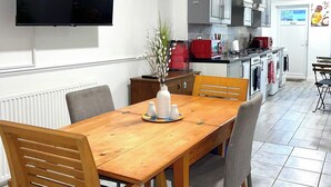Un comedor con una mesa y sillas de madera, un florero con ramas, y una cocina al fondo con electrodomésticos y armarios.