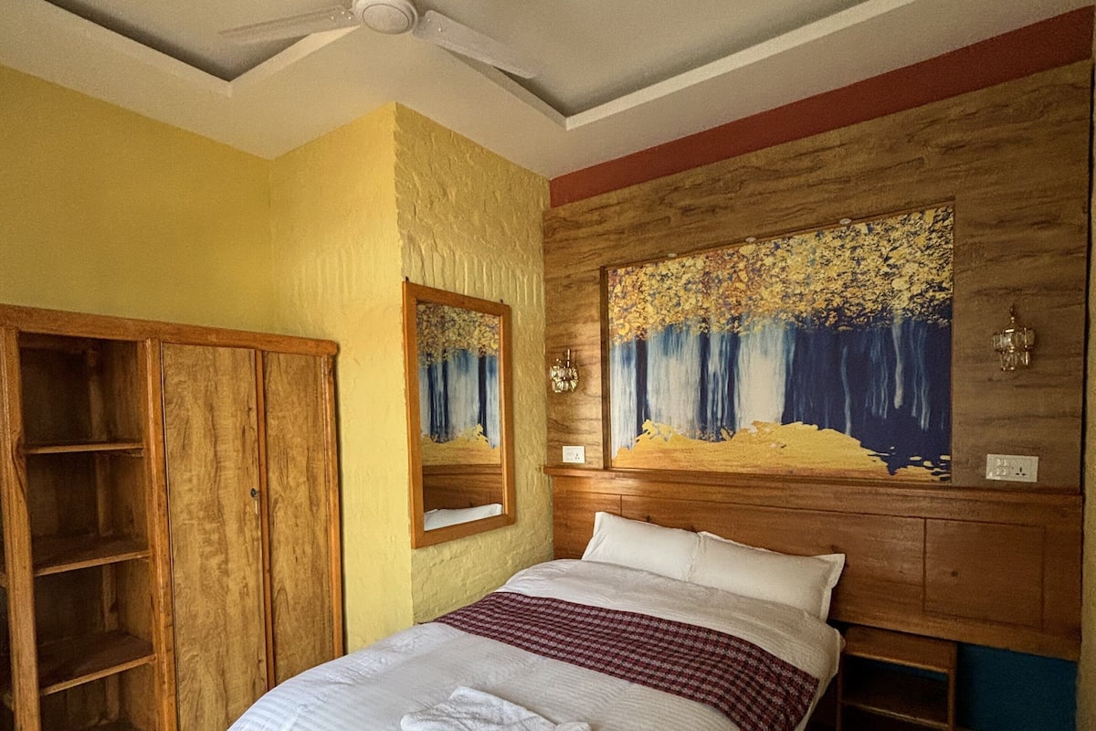 A bedroom with a bed, wooden wardrobe, ceiling fan, and a painting on the wall.