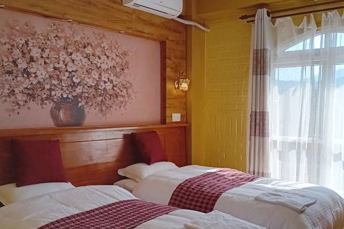 A hotel room with two beds, wooden headboard, a large floral artwork, and a window with sheer curtains.