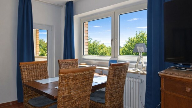 A room with a dining table, four wicker chairs, a television, and a window with a view of greenery and buildings.