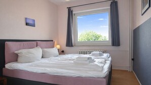 A neatly made bed with pillows and towels, a bedside lamp, a window with curtains, and a radiator.