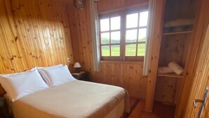 Un dormitorio de cabaña de madera con una cama, una mesita de noche, una ventana con vistas al exterior y un estante empotrado.