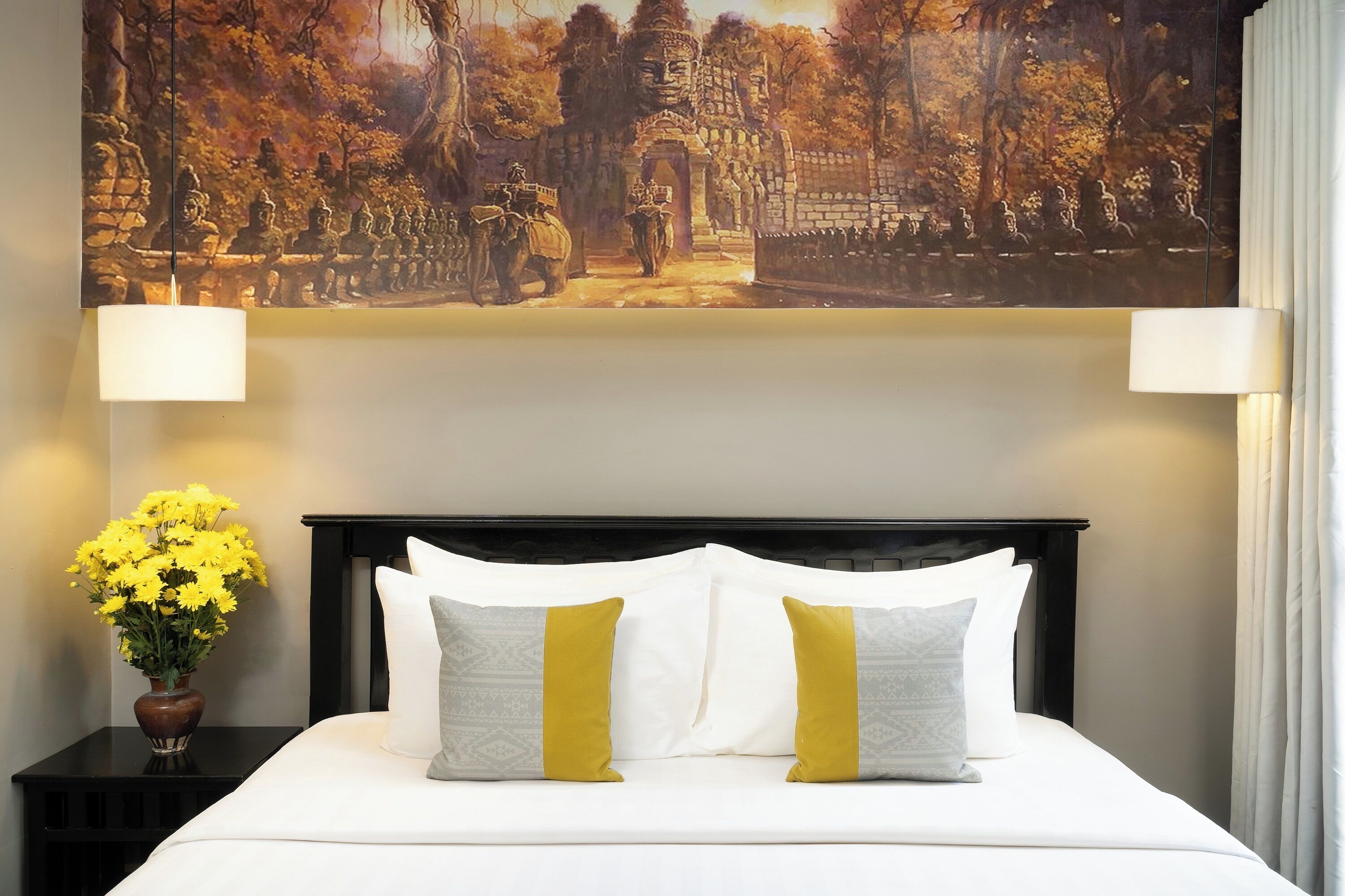 Una habitación de hotel con cama, ropa de cama blanca, un cuadro decorativo sobre la cama y un florero con flores amarillas en una mesita.