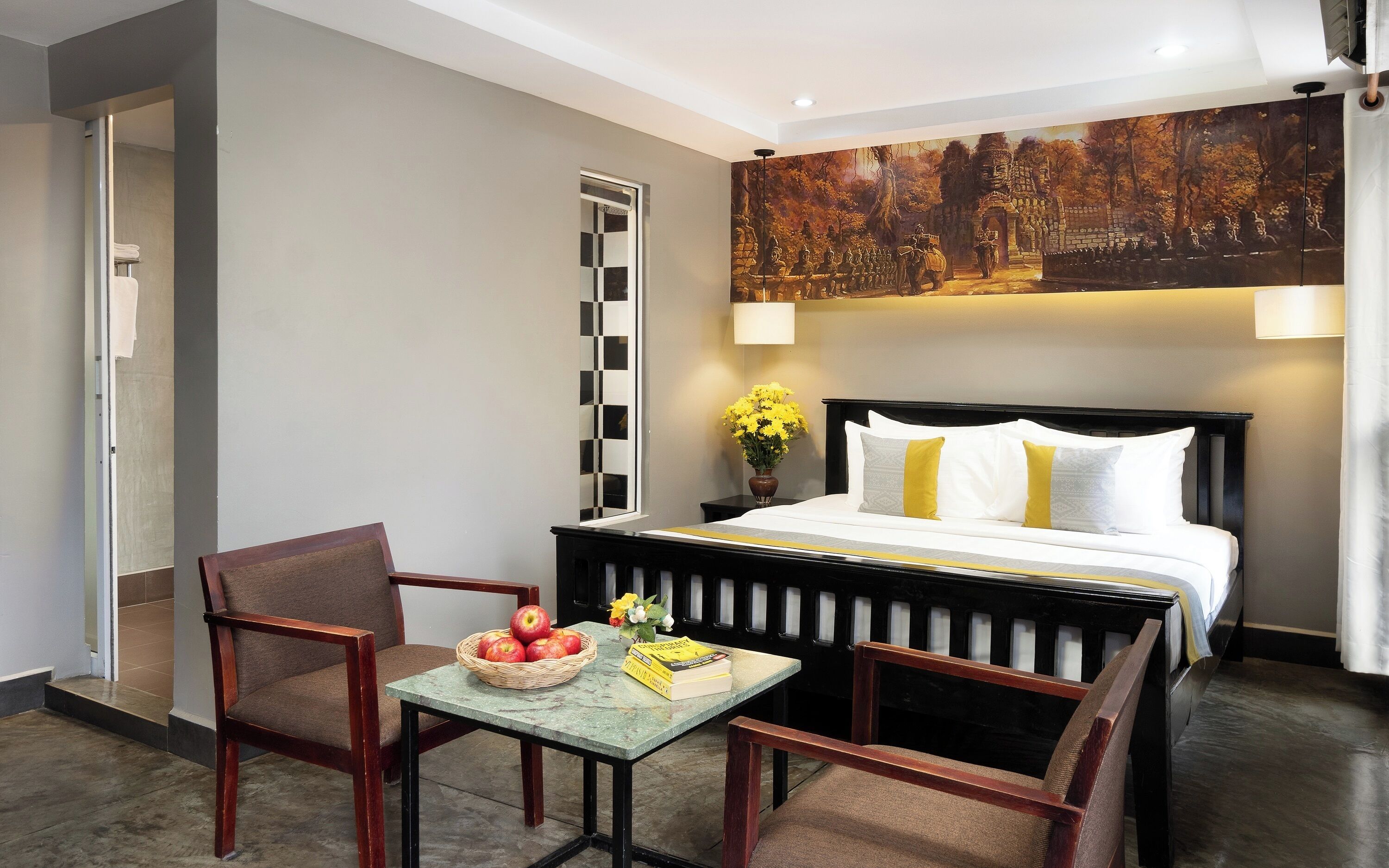 A hotel room with a bed, two chairs, a small table with apples and books, a painting on the wall, and a window with blinds.