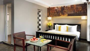 A hotel room with a bed, two chairs, a small table with apples and books, a painting on the wall, and a window with blinds.