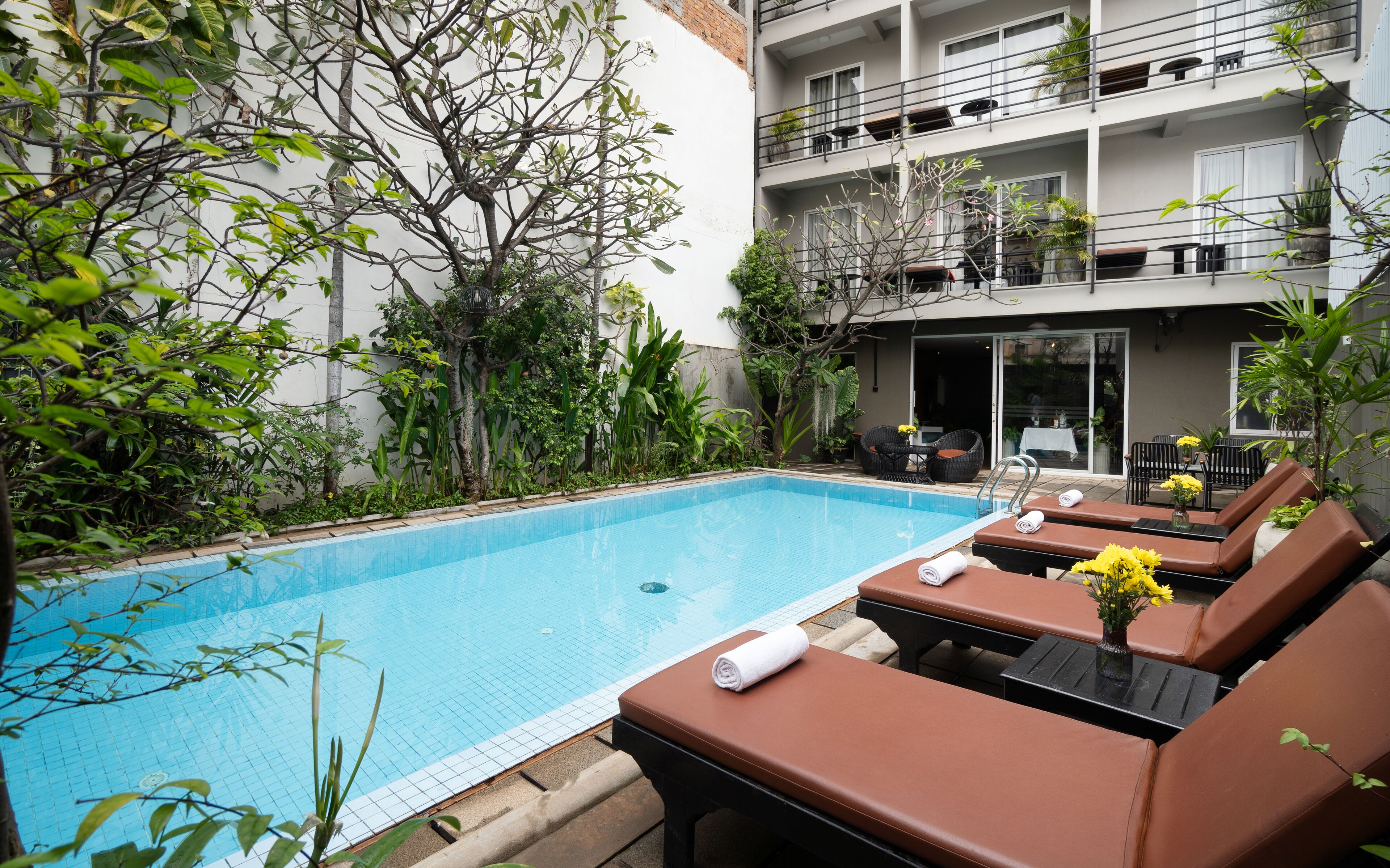 Un área junto a la piscina con sillones de descanso y una pequeña mesa, rodeada de vegetación y un edificio con balcones.
