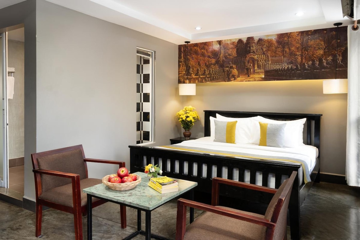 Una habitación de hotel con una cama, dos sillas, una mesita con fruta y libros, un cuadro en la pared y una puerta.