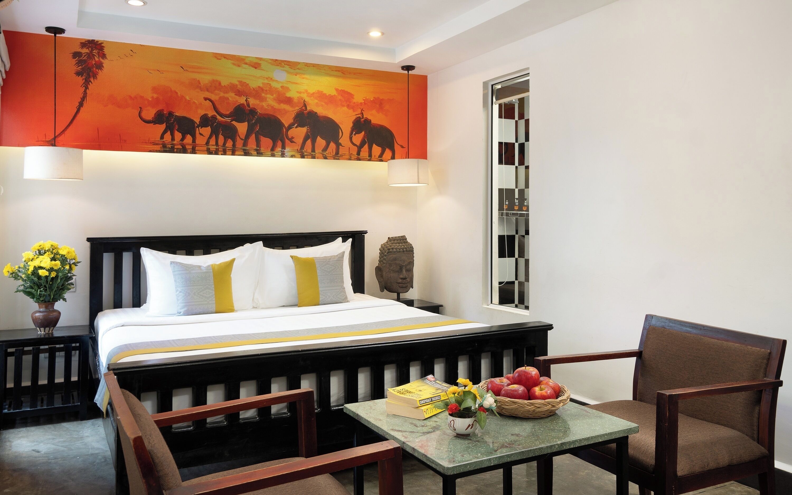 A hotel room with a large bed, a nightstand with a lamp, a small table with books and fruit, and a Buddha statue.