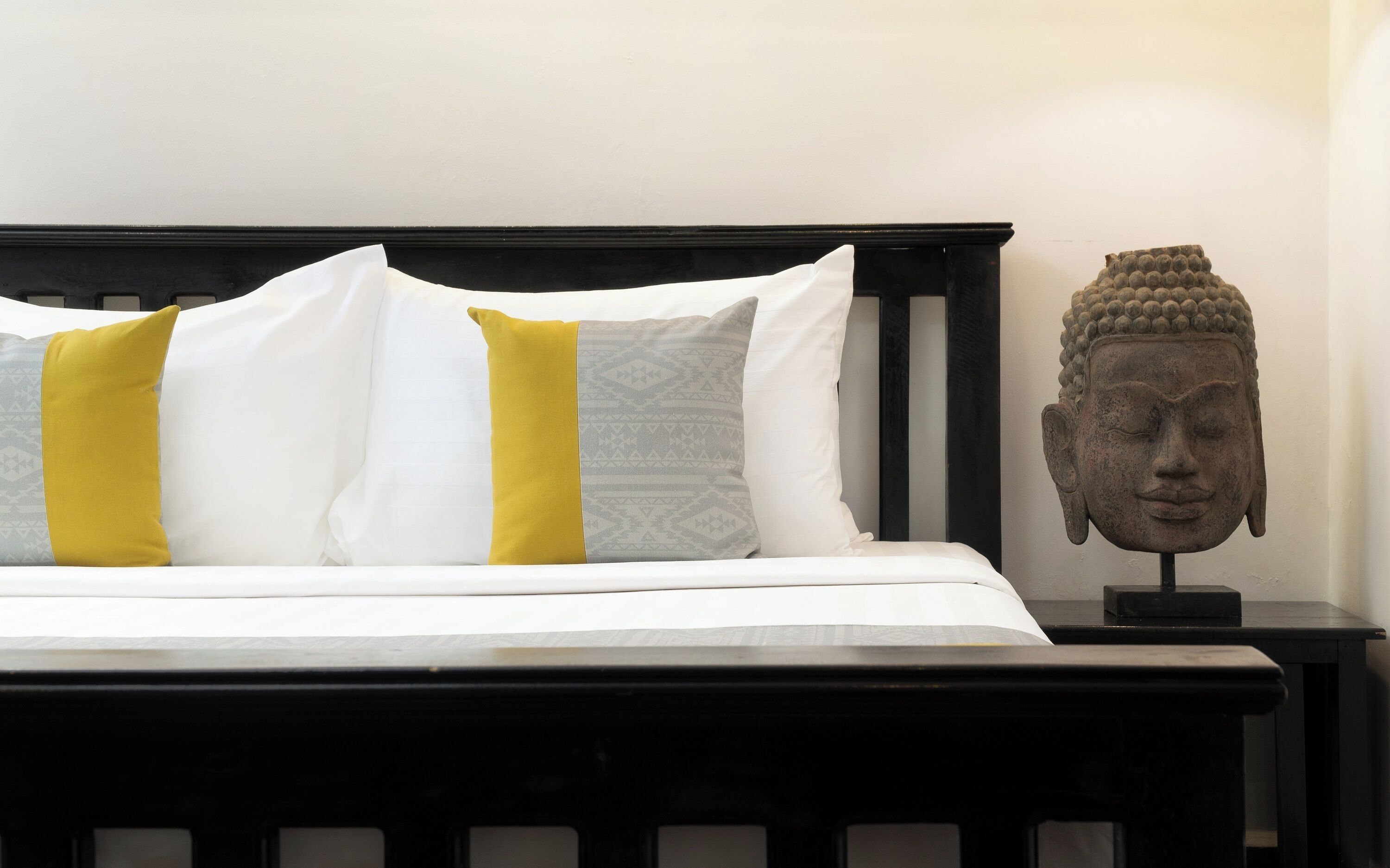 Una cama con almohadas blancas y amarillas, un cabecero de madera y una estatua de Buda sobre una mesita de noche.