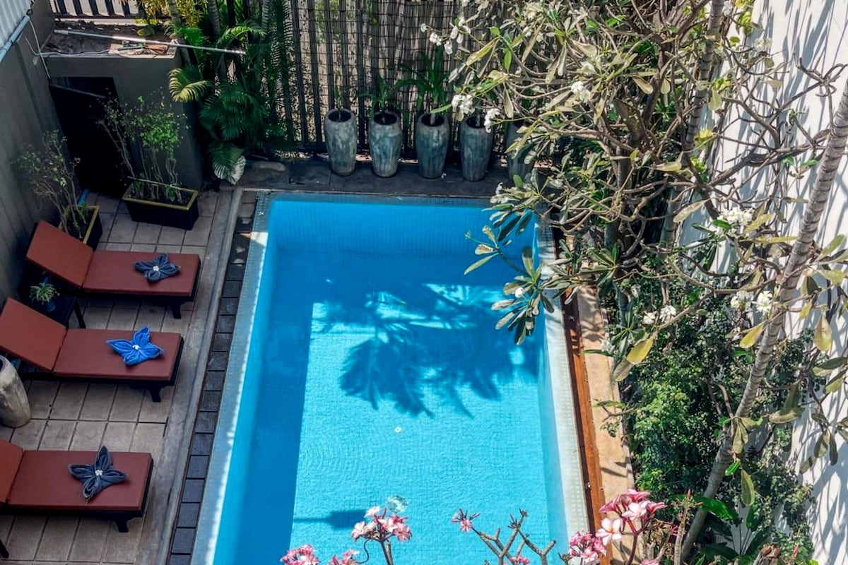 A rectangular pool with a tiled deck, surrounded by potted plants and a few lounge chairs.