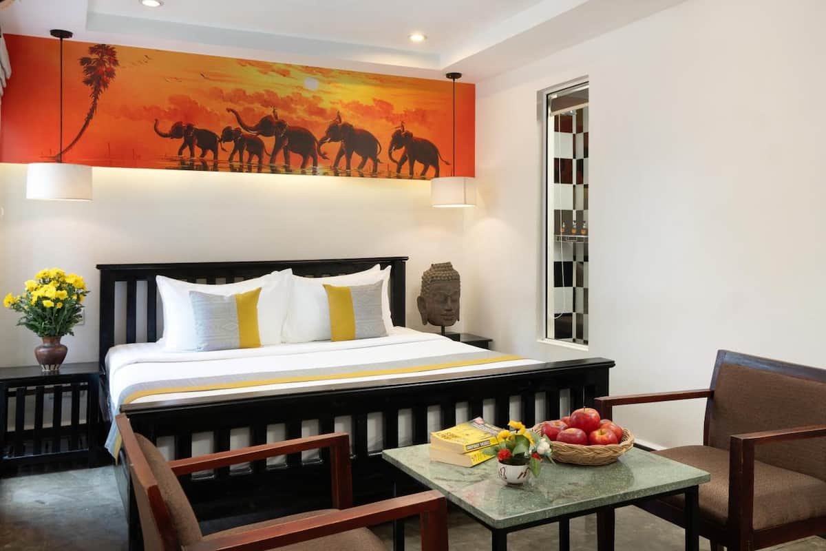 Een hotelkamer met een bed, een nachtkastje, een Boeddhabeeld, een schilderij van olifanten, een bloevas en een tafel met appels en boeken.