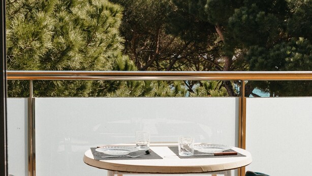 Mesa redonda con dos sillas, un vaso y un plato en un balcón con vistas a árboles.