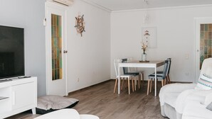 Una sala moderna con un televisor de pantalla plana, una zona de comedor con una mesa y sillas blancas, y un rincón de estar acogedor con un sofá.