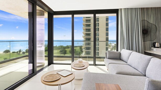 A modern living room with a grey sofa, two round tables with woven placemats, and a view of the ocean and a high-rise building. - B-702 Stunning ocean view (Cupecoy)