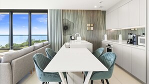 A modern kitchen with a central island, a dining area with a white table and teal chairs, and a large window offering a view of the sea. - B-702 Stunning ocean view (Cupecoy)