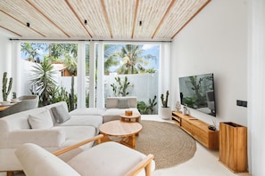 Uma sala de estar moderna com um sofá branco, uma mesa de centro redonda de madeira, uma televisão de tela plana e uma grande janela com vista para palmeiras.
