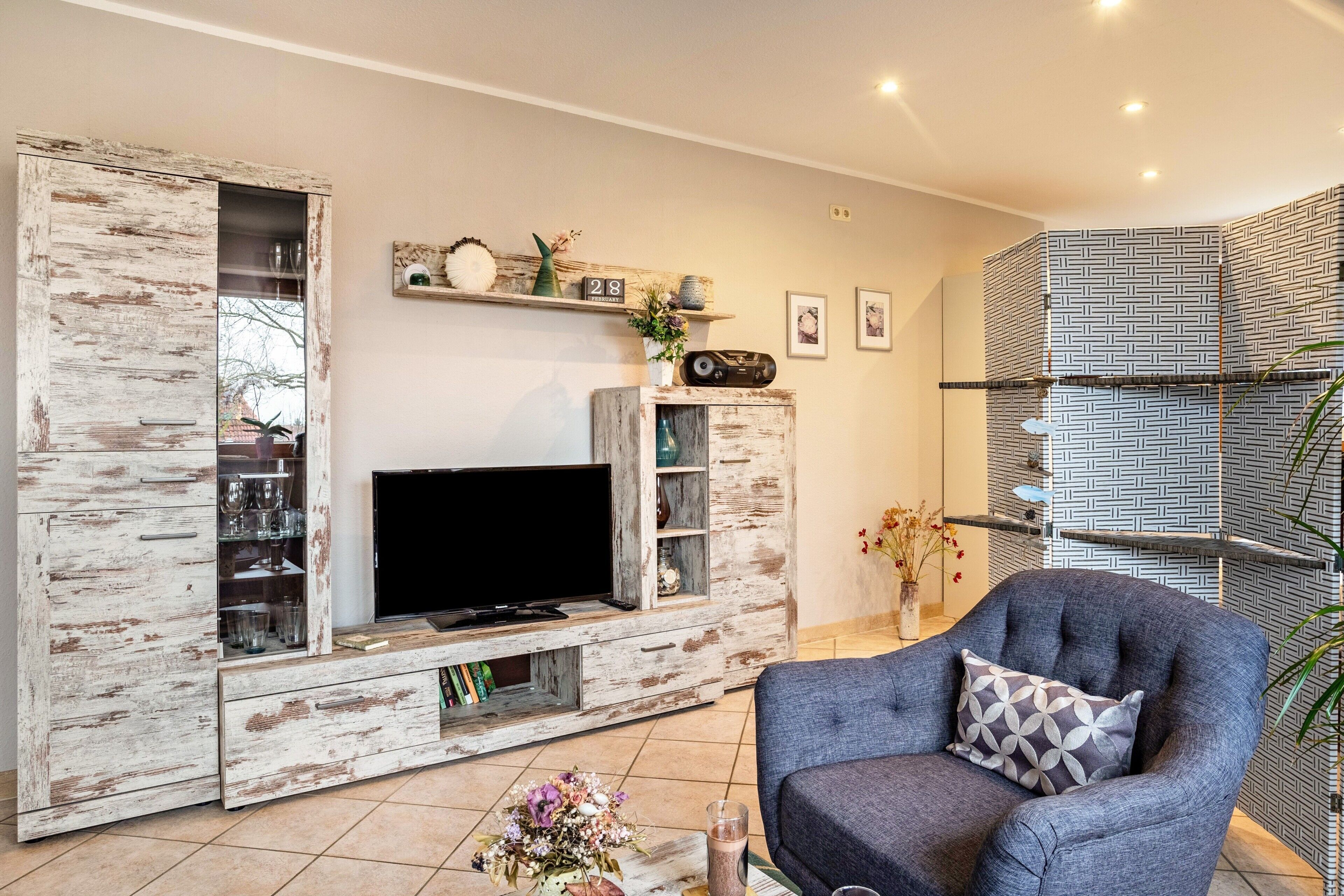 A living room with a TV, wooden entertainment unit, grey armchair, and patterned wall.