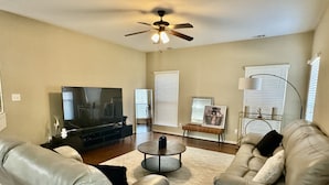 A living room with a large flat-screen TV, leather sofas, a coffee table, and a ceiling fan.