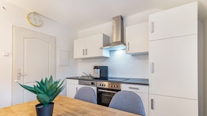 Une cuisine compacte avec des meubles blancs, une hotte de cuisinière en acier inoxydable, un four encastré et une table à manger avec deux chaises.