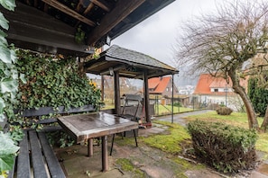 Un espace extérieur aménagé, abrité et doté de tables et de bancs, entouré de verdure et offrant une vue sur un quartier résidentiel.